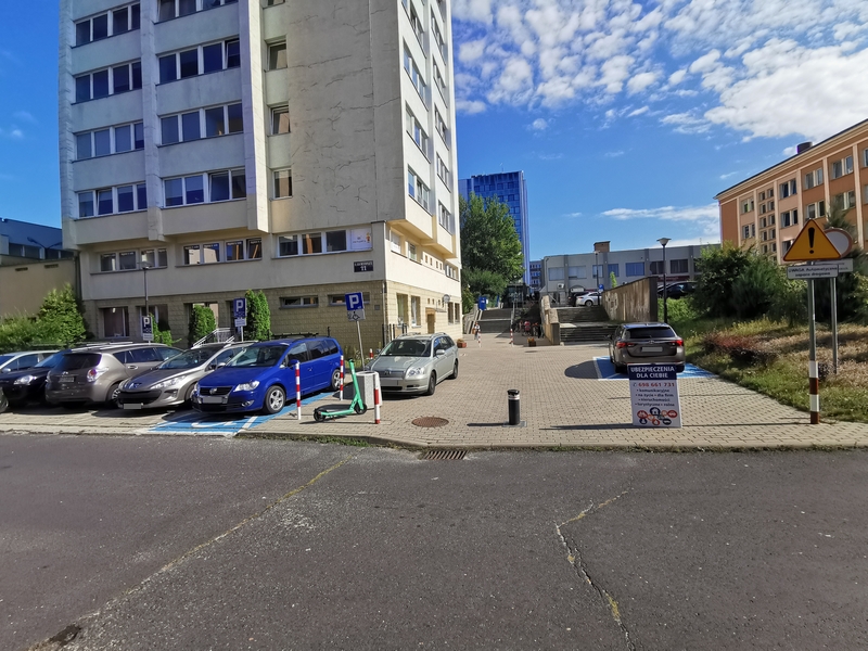 Zdjęcie przedstawia parking z samochodami przy biurowcu, w którym znajduje się Inspektorat, na którym widać dwa oznaczone miejsca do parkowania dla osób z niepełnosprawnościami