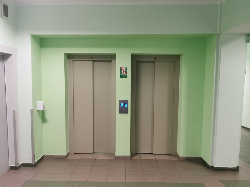 Zdjęcie przedstawia hol biurowca oraz drzwi dwóch wind dostosowanych dla osób ze specjalnymi potrzebami na tle zielonej ściany