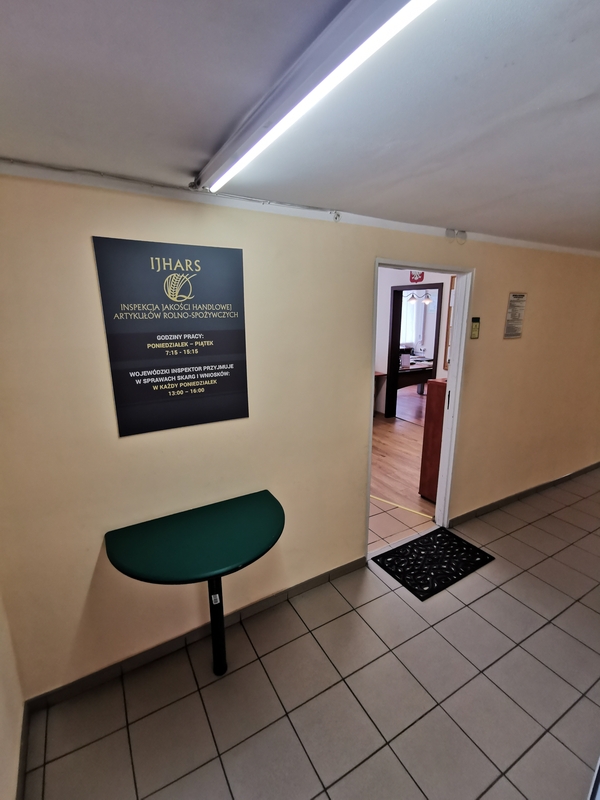 Zdjęcie przedstawia wejście do sekretariatu Inspektoratu. Na ścianie widzimy zieloną tablicę informacyjną oraz otwarte drzwi. 